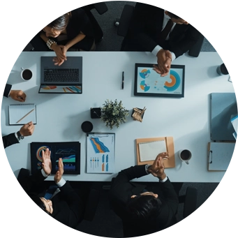 Veri grafiklerini ve tablolarını dijital tabletlerde tartışan insanların bir masa etrafında toplandığı bir iş toplantısının üstten görünümü.